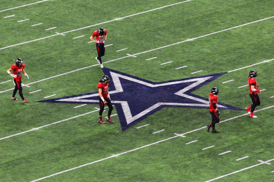 Five Varsity players walk across the Cowboys logo on field.